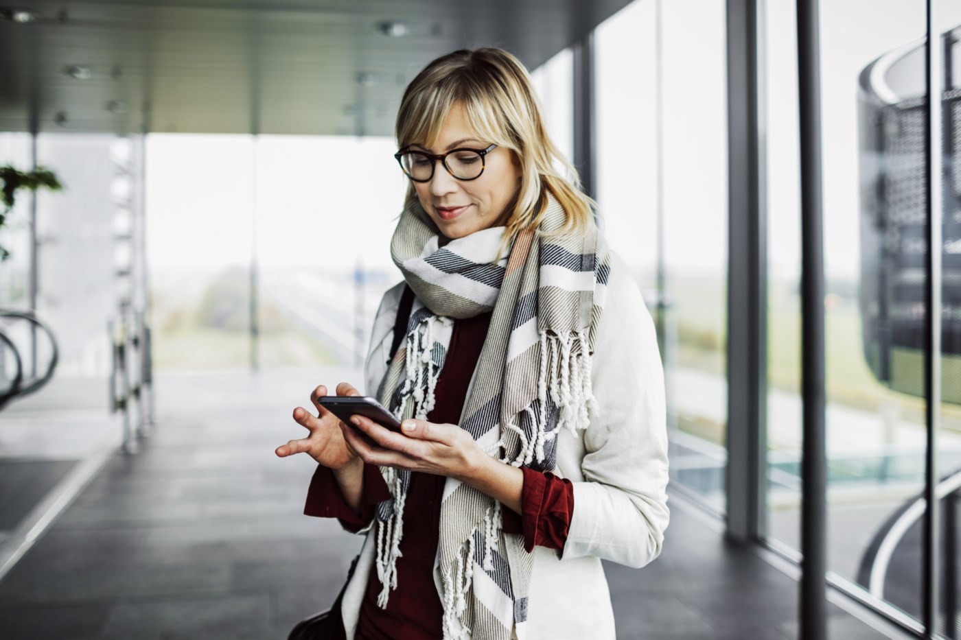 Velkledd dame ser på mobilen sin på en flyplass. Hun smiler lett.